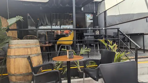 La terraza de día, con un barril, sillas negras y plantas junto a la barandilla