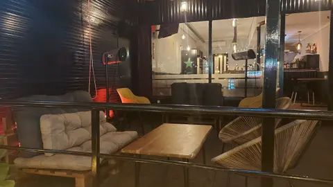 La terraza de noche: sofás de madera con cojines, mesas bajas y la pared de chapa negra con luz roja